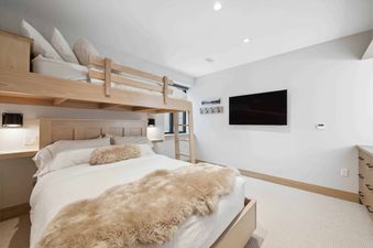 A modern bedroom at One Riverfront Townhome 05 features a light wood twin-over-queen bunk bed with white bedding, a faux fur throw, a wall-mounted TV, and a window.