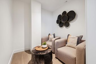 Two tan armchairs with pillows face each other in a room with a dark wood floor, a white wall, and a decorative wall piece made of black circular plates with gold trim.