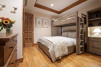 A bunk bed bedroom with light wooden furniture, a dresser with flowers, and a built-in cabinet with a lamp, featuring hardwood floors and a decorative ceiling.
