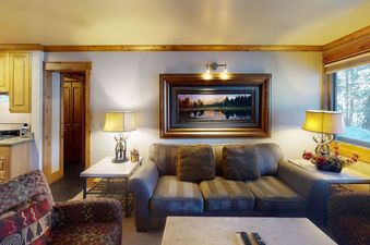 Manor Vail 173 living room with a couch facing a large framed painting and a table with a lamp. There are floor lamps on either side of the couch and a window showing a view of the snowy trees.