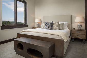 Lumiere Three Bedroom Luxury bedroom features a king size bed with white linens, decorative pillows, a wood frame, and a throw blanket. The bedroom also includes a small side table with a lamp and a small bench at the foot of the bed. The view from the wi