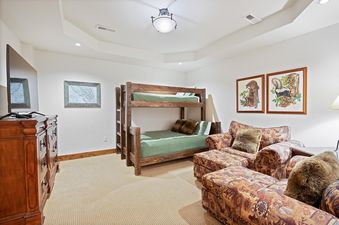 The bunk room at Whitefish Ski Haus features a rustic wooden bunk bed with green bedding, a patterned sectional sofa, a wooden dresser with a television, and nature-themed artwork, all set under a tray ceiling with warm lighting.