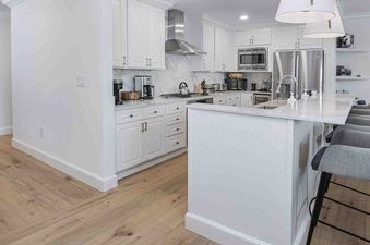 A modern kitchen with white cabinets, stainless steel appliances, a kitchen island with bar stools, and light wood flooring.