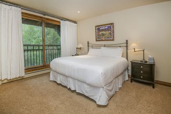 A cozy bedroom at Pines Townhome 10 with a king bed, nightstands, lamps, artwork, and a balcony with an exterior view of lush greenery.