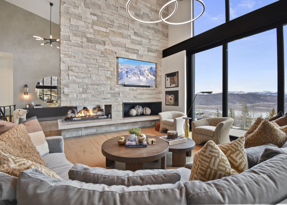 A spacious and luxurious living room with a large stone fireplace, a cozy sofa, two round coffee tables, and floor-to-ceiling windows offering a panoramic view of snowy mountains and a valley.