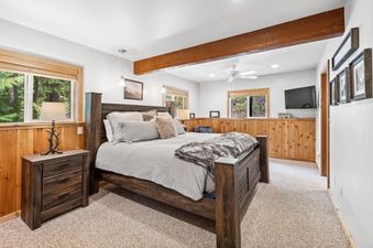 A rustic bedroom with a large wooden bed, soft bedding, and wood-paneled walls, featuring a view of lush greenery outside the windows.