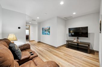 A cozy living room in Pioneer Peak features a brown leather sectional sofa with cup holders, a wall-mounted flat-screen TV over a dark wood console table, and nature-themed art on the light-colored walls.