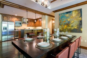 A dining table set for six with wine glasses, plates, and silverware, and a large abstract painting on the wall behind it. The table is made of dark wood and has a polished finish, and the chairs are upholstered in red leather.