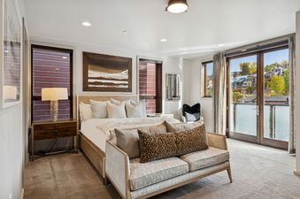 A modern primary bedroom at Main Street Modern, featuring a king-sized bed with white bedding, a bedside table with a lamp, a long settee, and large windows offering an exterior view.