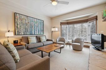 The living room of The Charter D340 features a comfortable gray sectional, a wooden coffee table, and a large bay window overlooking snow-covered trees.