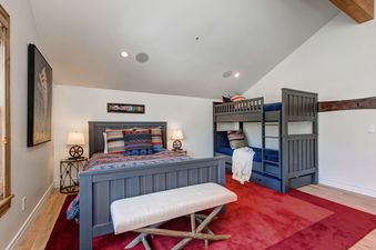 A bedroom with a large bed, a bunk bed, a white bench, and a red rug.