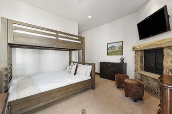 A cozy bunk room featuring a wooden bunk bed with white bedding, a dark wood dresser, a stone fireplace, and a wall-mounted TV.