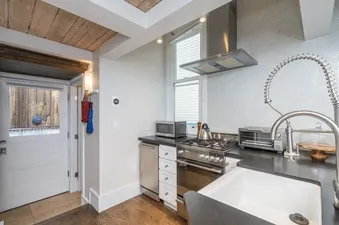 A modern kitchen at The Hummingbird House with stainless steel appliances, dark countertops, a white farmhouse sink, and a rustic wooden beamed ceiling.