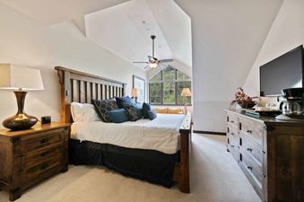 A serene bedroom at St James Place 437 featuring a large wooden bed with blue pillows, a wooden dresser with a TV and coffee maker, and a gabled window overlooking trees.