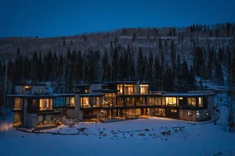 White Pine Canyon is a luxurious, modern home nestled in the snowy woods. The exterior features large windows that provide stunning views of the surrounding nature, with a snowy yard and trees in the background.