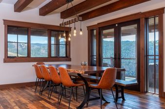 A rustic modern dining area at Granby Ranch Lodge featuring a live-edge table, orange-brown leather chairs, and large windows looking out to forested hills.