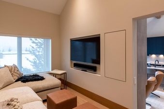 A bright and modern lounge area featuring a large sectional sofa, a wall-mounted TV, and a view of a snowy landscape through a large window, with a partial view of a modern kitchen in the background.