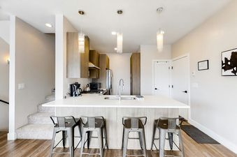 A modern kitchen with a white island, four bar stools, light wood cabinets, and stainless steel appliances.