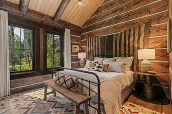 A rustic bedroom with log walls, wood beams, a metal-frame bed, and large windows looking out to trees.