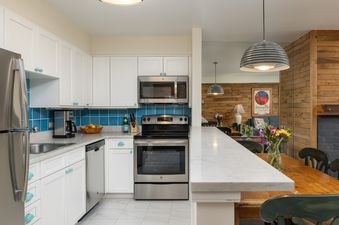 The contemporary kitchen in Montaneros 311 features white cabinetry, stainless steel appliances, a vibrant blue tiled backsplash, and a marble breakfast bar, seamlessly connecting to a dining area with a rustic wooden table and a cozy lounge.