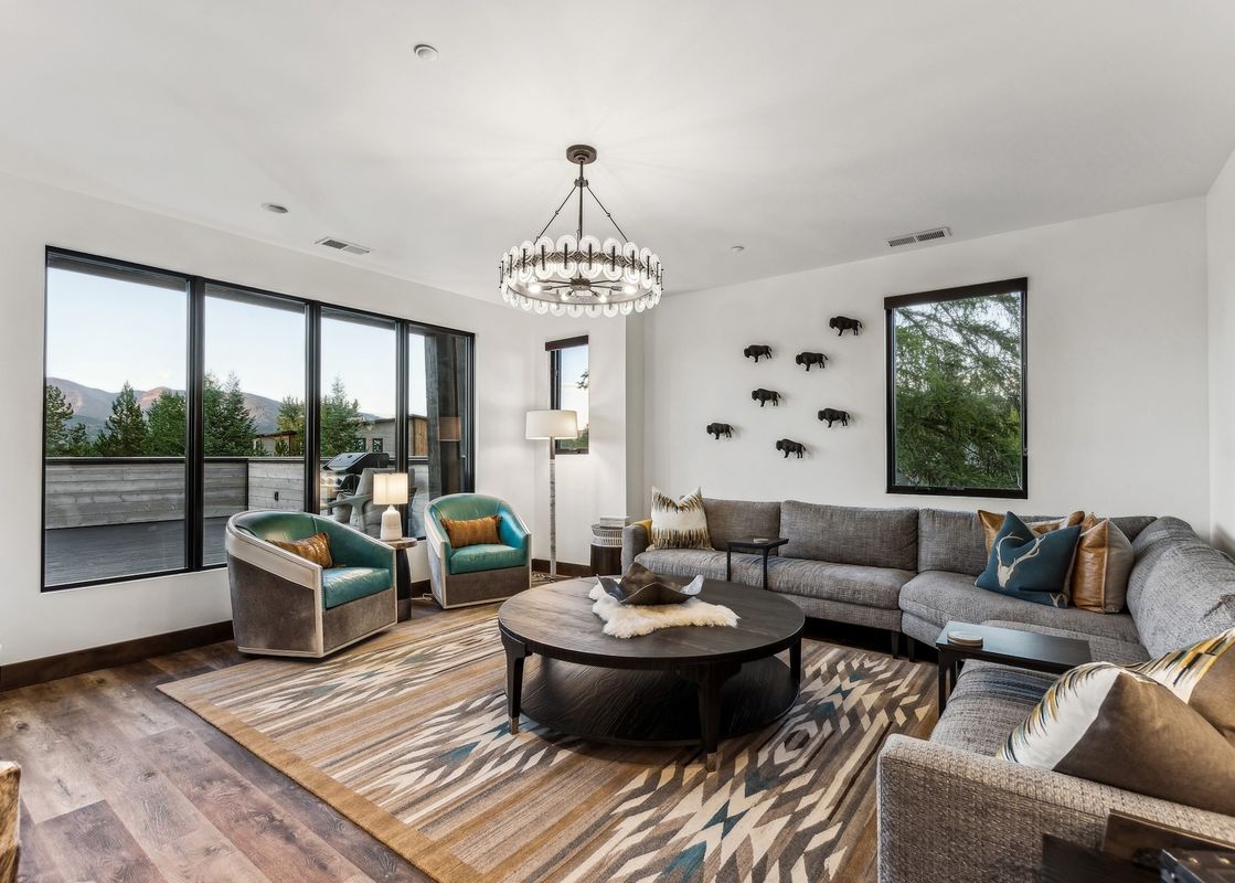 A spacious and stylish living room with a large sectional sofa, two teal armchairs, a round coffee table, and large windows offering mountain views.