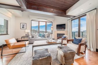 The living room of Lumiere 2 Bedroom Suite, with a large window offering views of the surrounding mountains, a comfortable sofa and chairs, a coffee table, and a fireplace.