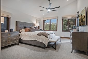A spacious bedroom at Antlers Estate featuring a large bed with a gray upholstered headboard, multiple pillows, and a white duvet, with two large windows offering a scenic view of a snowy mountain.