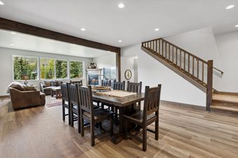 A wide view of the open-concept living and dining area at Pioneer Peak, featuring a large wooden dining table, a cozy living room with a fireplace, and expansive windows offering views of the forest.