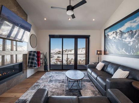 A spacious and elegantly furnished living room in the Penthouse in the Pines features a cozy fireplace, comfortable sofas, and a large window offering panoramic views of a snowy mountain landscape.