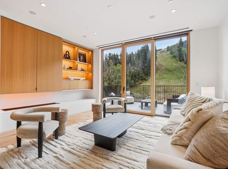The Founders Place 2401 living area features a cream sofa, modern armchairs, and a sleek coffee table, with a built-in wooden cabinet and large glass doors leading to a balcony with mountain views.