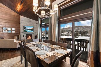 A luxurious dining room at Lakeside 1629 features a large wooden table set for dinner, a stone fireplace, and floor-to-ceiling windows offering a panoramic view of a snow-covered landscape.