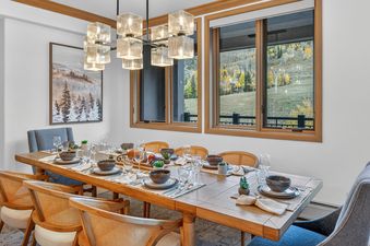 A beautifully set dining table with rattan and upholstered chairs, lit by a modern chandelier, offers views of an autumn mountainside.