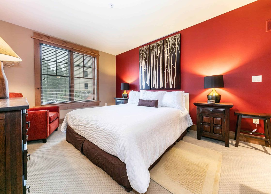A bedroom with a white duvet and brown bed skirt, a brown end table with a lamp and a red chair in the corner.