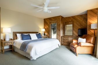 The luxurious bedroom at 88 Ridge Point features a comfortable king-sized bed with white linens and blue accents, two elegant nightstands with lamps, a cozy brown leather armchair, and a large flat-screen TV.