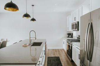 A modern kitchen with a large island and black pendant lights features white cabinetry, stainless steel appliances, and wood flooring.