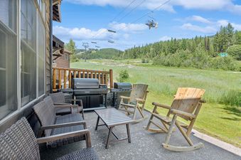 A patio at Slopeside 117 with wicker chairs, a coffee table, and two BBQs, offering views of green ski slopes and a ski lift under a blue sky.