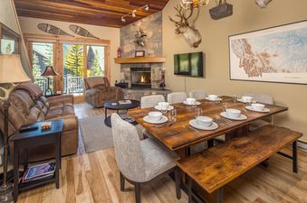 A spacious living and dining area with a stone fireplace, leather furniture, a large wooden dining table, and rustic decor, all bathed in warm lighting.
