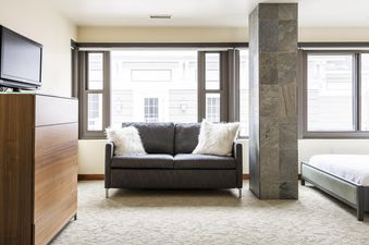 A cozy living space in Lowell 401A featuring a dark gray sofa with white fluffy pillows, a wooden dresser with a TV, and large windows offering an exterior view.