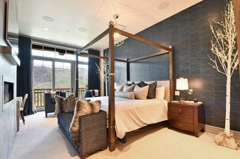 A king bedroom features a four-poster bed, a dark wood nightstand, a modern lamp, and a textured blue accent wall, with floor-to-ceiling windows offering a mountain view.