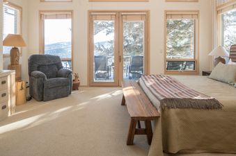 A cozy bedroom with a comfortable bed, recliner, and a balcony with outdoor chairs, offering scenic views of snow-covered hills and pine trees.