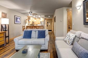 The spacious open-concept living area at Snow Flower 132 Park City features comfortable light-colored sofas, a light-green coffee table, and an adjacent kitchen with light wood cabinetry and stainless steel appliances.