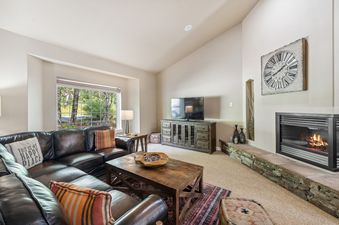 A cozy living room at Bitterroot Escape featuring a brown leather sectional sofa, a wooden coffee table, a flat-screen TV on a console, and a fireplace with a stone base.