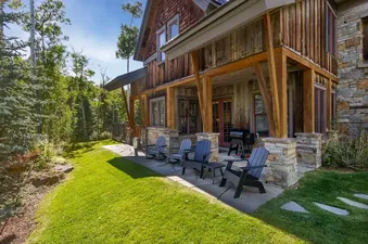 An inviting outdoor patio at the Golden Knoll featuring wooden Adirondack chairs, a BBQ grill, and a rustic wooden and stone exterior, surrounded by lush green lawn and trees under a clear blue sky.