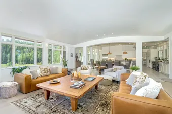 A spacious and inviting living room at Turkey Creek with brown leather sofas, light gray armchairs, a rustic wooden coffee table, and large windows overlooking a lush green forest.