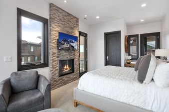 A luxurious bedroom at Juniper 901 features a plush bed, a modern fireplace with a TV mounted above, a comfortable armchair, and a window with a view of nearby buildings.