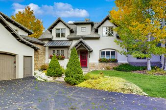 A charming home with a red front door, light stucco walls, and a stone foundation, set against a blue sky with autumn trees and a neatly manicured lawn.