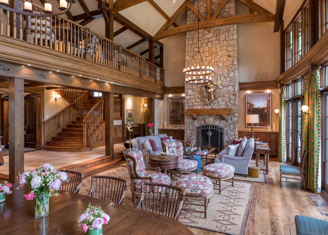 A rustic living room with a stone fireplace, large windows, and a wooden staircase leading to a balcony.