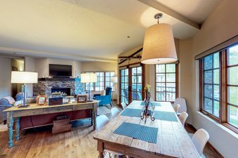 The dining area of Westermere 310 features a rustic wooden table with blue placemats, a chandelier, and a view of the mountains through the windows.
