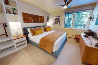 A bedroom with a king-sized bed, a large window with a view of a forest, a TV, and a dresser with a marble top.