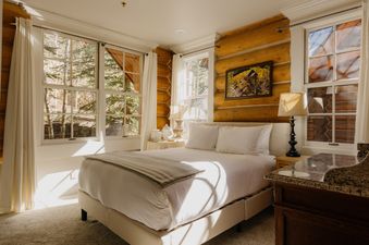 A bright bedroom in the Summit Cabin at See Forever with a bed, large windows looking out to trees, and log walls.